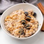 A bowl of overnight oats topped with blueberries, chia seeds, and a PB drizzle, accompanied by cinnamon sticks and a gold spoon.