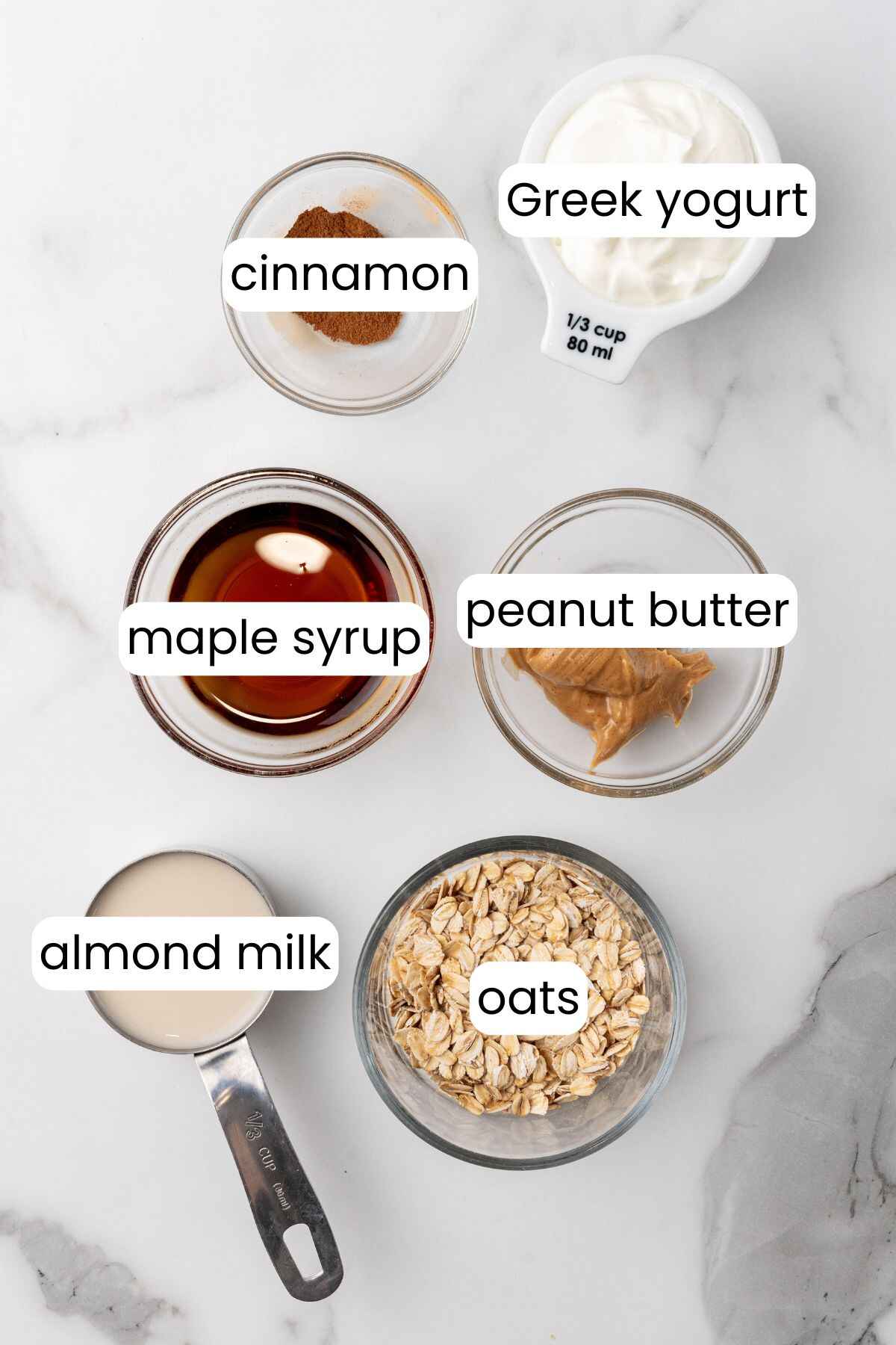 Labeled ingredients on a marble surface: cinnamon in a bowl, Greek yogurt in a cup, chia seeds sprinkled next to PB Overnight Oats, maple syrup in a bowl, peanut butter on a spoon, almond milk in a measuring cup, and oats in a bowl.