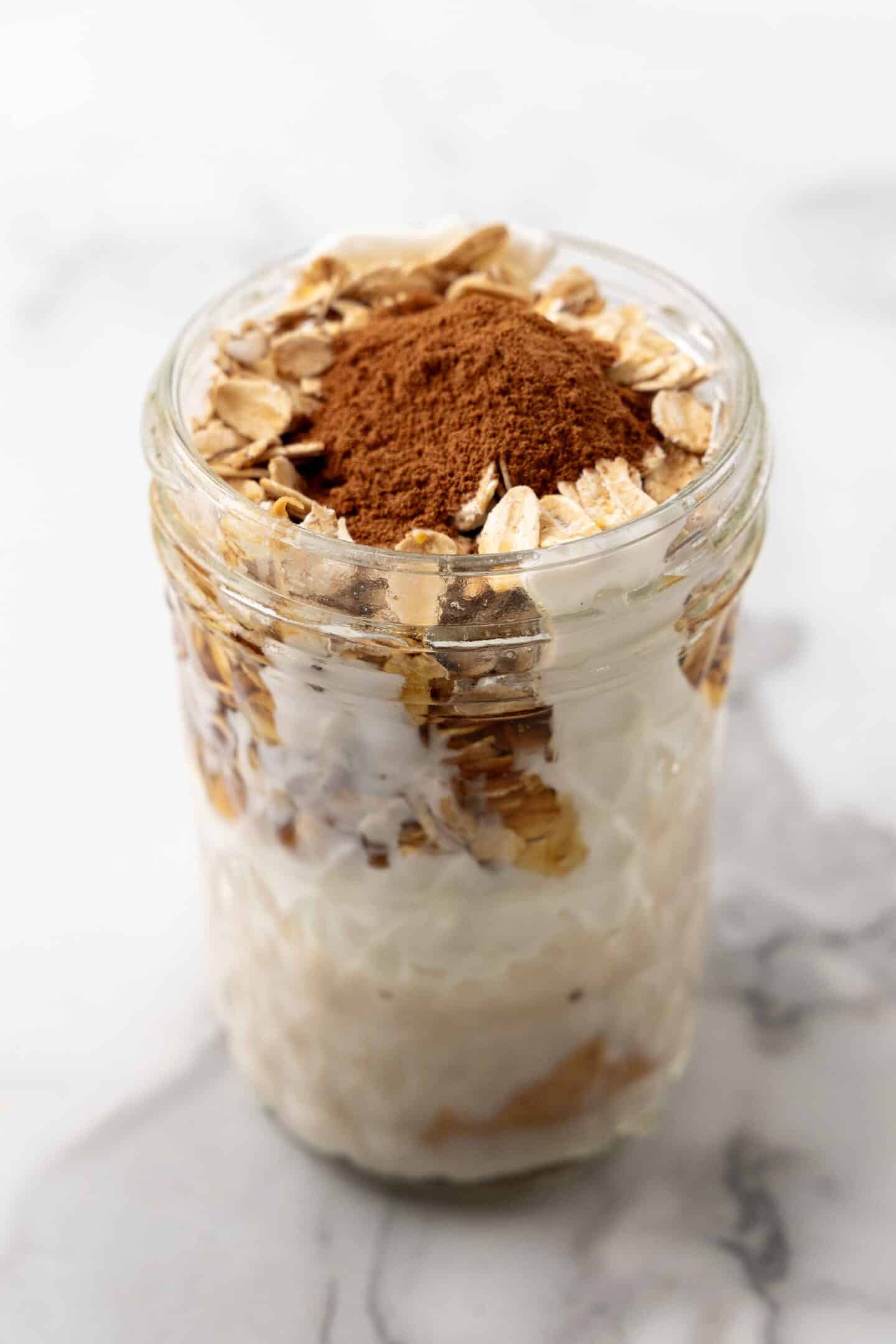 A jar filled with delicious overnight oats and creamy yogurt, elegantly layered and topped with a sprinkle of cocoa powder, rests on a marble surface.