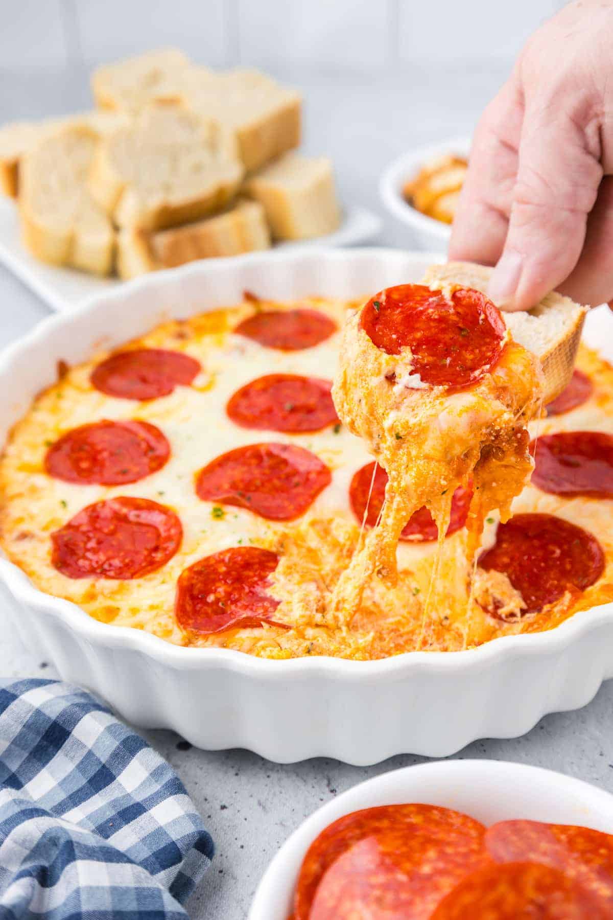 A hand dips bread into a pepperoni-topped cheese dip in a white dish, perfect for Super Bowl snacks, with sliced bread and a checkered napkin nearby.