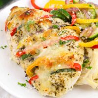 Grilled Primavera Stuffed Chicken breast with melted cheese and herbs, served with sliced bell peppers, zucchini, and onions on a bed of pasta on a white plate.