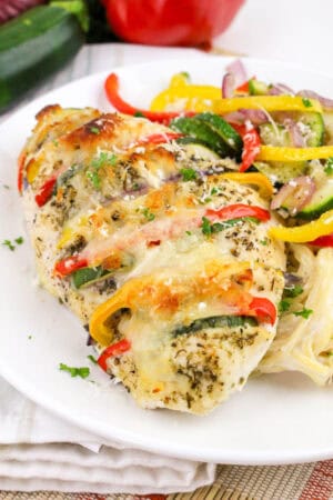 Grilled Primavera Stuffed Chicken breast with melted cheese and herbs, served with sliced bell peppers, zucchini, and onions on a bed of pasta on a white plate.