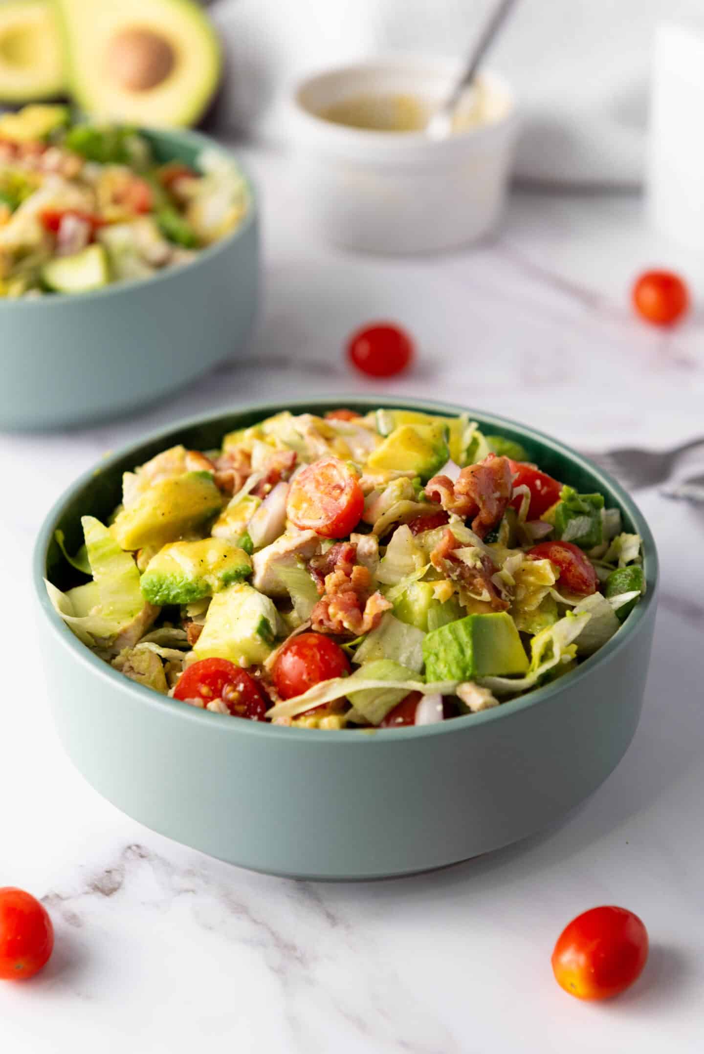 A bowl of chicken chopped salad with lettuce, avocado, cherry tomatoes, and bacon rests on a marble surface. Another bowl and cherry tomatoes are in the background. Try drizzling honey mustard dressing for an extra flavor twist to this delightful salad recipe.