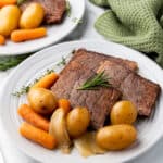 Two plates with slices of succulent rump roast, accompanied by tender baby carrots, potatoes, and onion, garnished with rosemary and thyme. The mouthwatering dish, reminiscent of a perfect Crock Pot meal, rests on a marble surface. A green napkin adds a touch of color to the background.