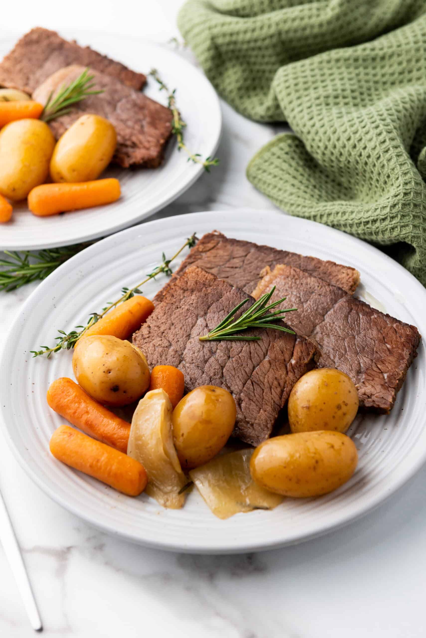 Two plates with slices of succulent rump roast, accompanied by tender baby carrots, potatoes, and onion, garnished with rosemary and thyme. The mouthwatering dish, reminiscent of a perfect Crock Pot meal, rests on a marble surface. A green napkin adds a touch of color to the background.