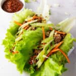 Korean lettuce wraps filled with Korean ground beef, shredded carrots, and green onions, garnished with sesame seeds. A bowl of dipping sauce is visible in the background.