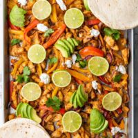 A vibrant sheet pan brimming with chicken fajitas, featuring a colorful medley of sliced bell peppers, onions, lime wedges, avocado slices, and fresh cilantro. Soft flour tortillas are artfully arranged around the pan for an inviting meal.