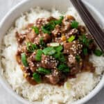 A bowl of Mongolian Beef over rice, featuring tender, saucy beef garnished with chopped green onions and sesame seeds, all cooked to perfection in a slow cooker. Chopsticks rest elegantly on the bowl's edge.