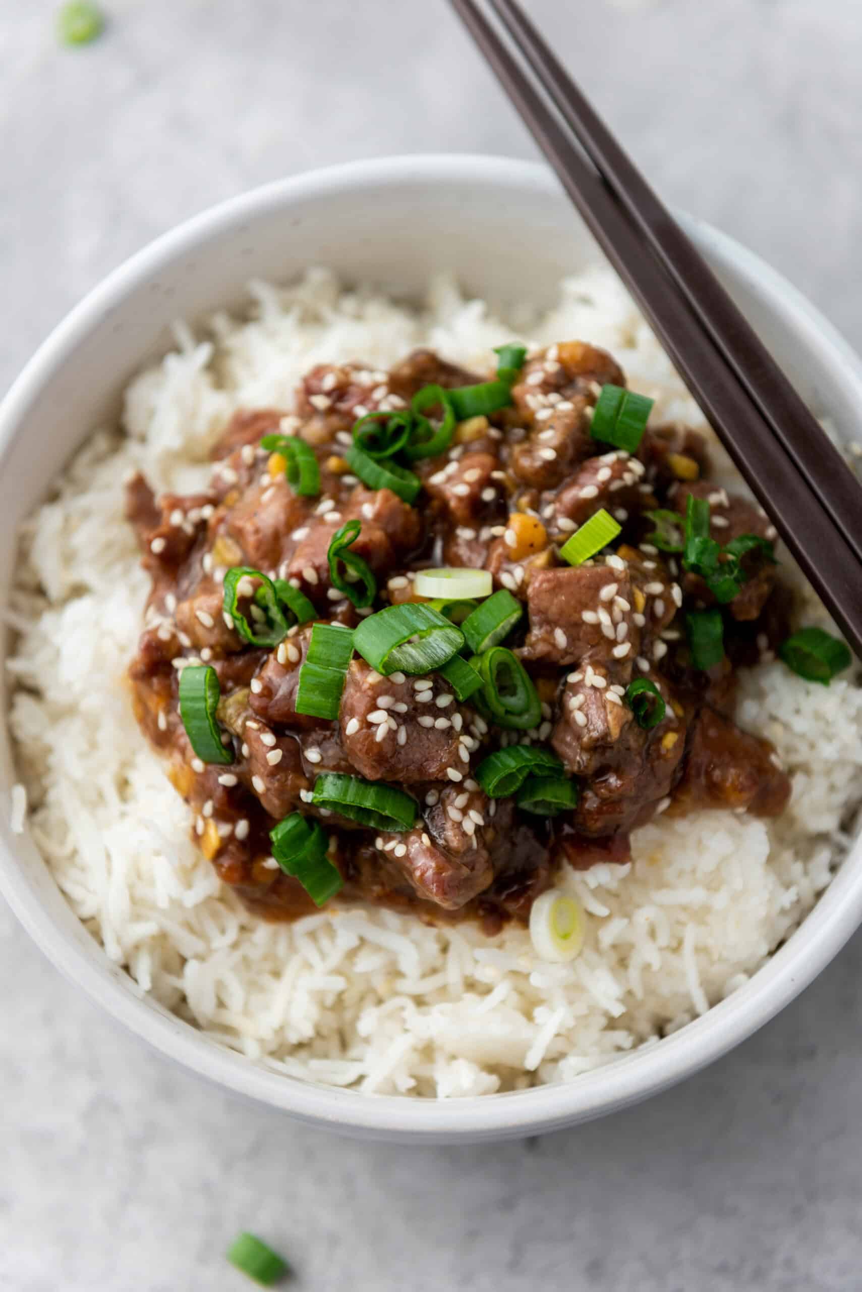 A bowl of Mongolian Beef over rice, featuring tender, saucy beef garnished with chopped green onions and sesame seeds, all cooked to perfection in a slow cooker. Chopsticks rest elegantly on the bowl's edge.