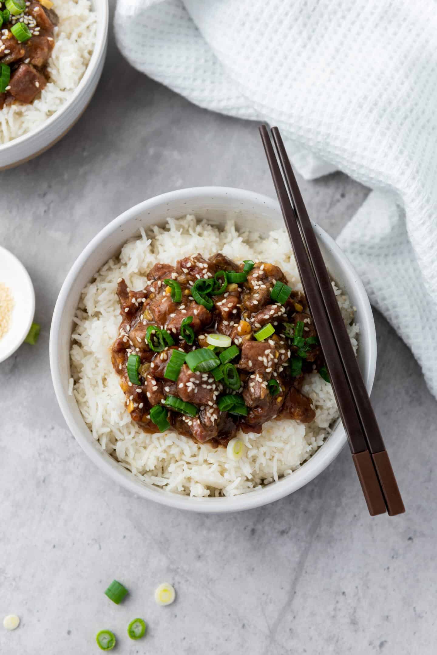 A bowl of tender Mongolian beef atop rice, garnished with chopped green onions and sesame seeds, with chopsticks resting on the side.