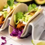Two tacos with grilled meat and blackened tilapia, sliced onion, cilantro, and green sauce on a taco holder. Red cabbage and a lime wedge are nearby on a tiled surface. An avocado sits in the background alongside a zesty avocado cilantro sauce.