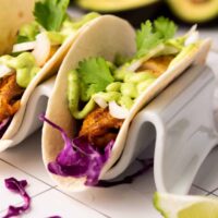 Two tacos with grilled meat and blackened tilapia, sliced onion, cilantro, and green sauce on a taco holder. Red cabbage and a lime wedge are nearby on a tiled surface. An avocado sits in the background alongside a zesty avocado cilantro sauce.