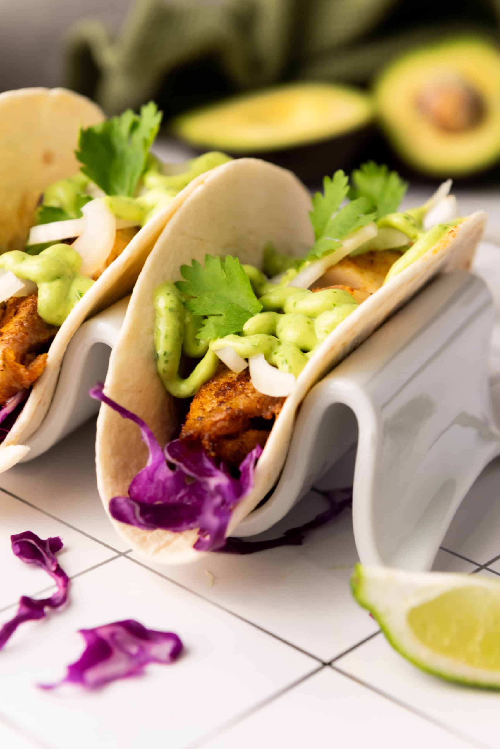 Two tacos with grilled meat and blackened tilapia, sliced onion, cilantro, and green sauce on a taco holder. Red cabbage and a lime wedge are nearby on a tiled surface. An avocado sits in the background alongside a zesty avocado cilantro sauce.