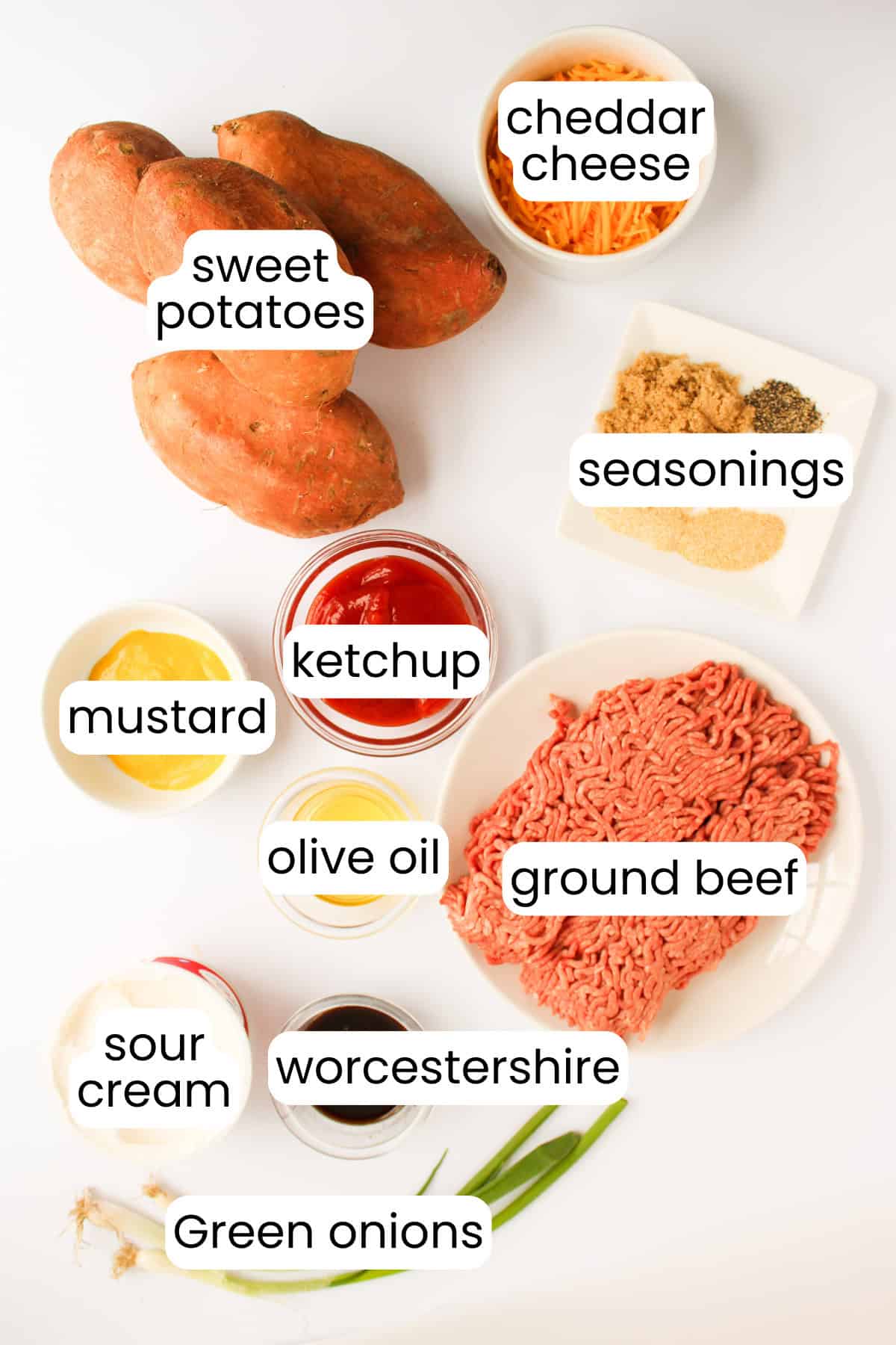 Ingredients on a white surface include sweet potatoes, cheddar cheese, seasonings, ground beef for Sloppy Joes, green onions, ketchup, mustard, sour cream, olive oil, and Worcestershire sauce.