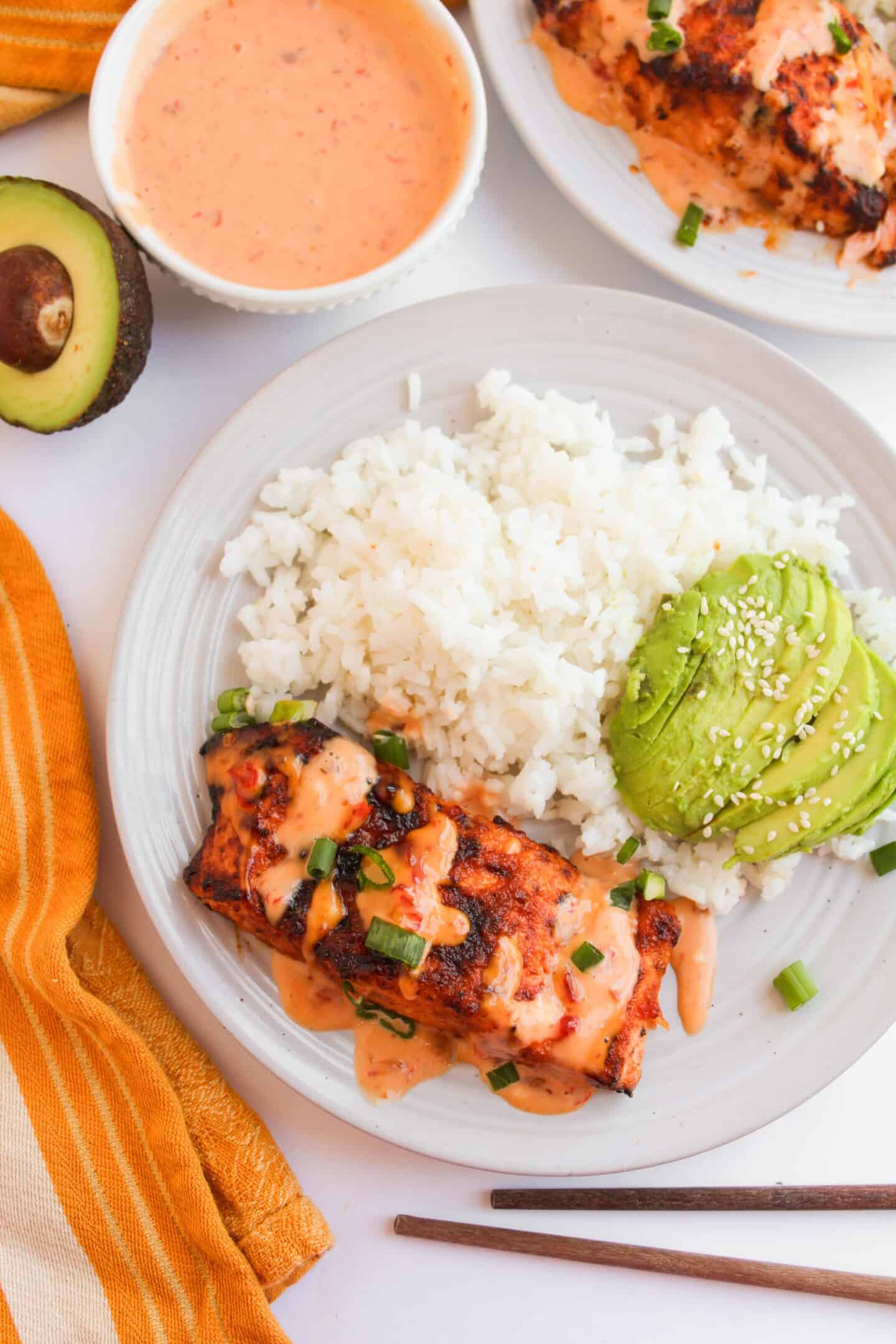Plate of Bang Bang Salmon with spicy sauce, white rice, and sliced avocado, garnished with sesame seeds and green onions. Chopsticks and an avocado are nearby.