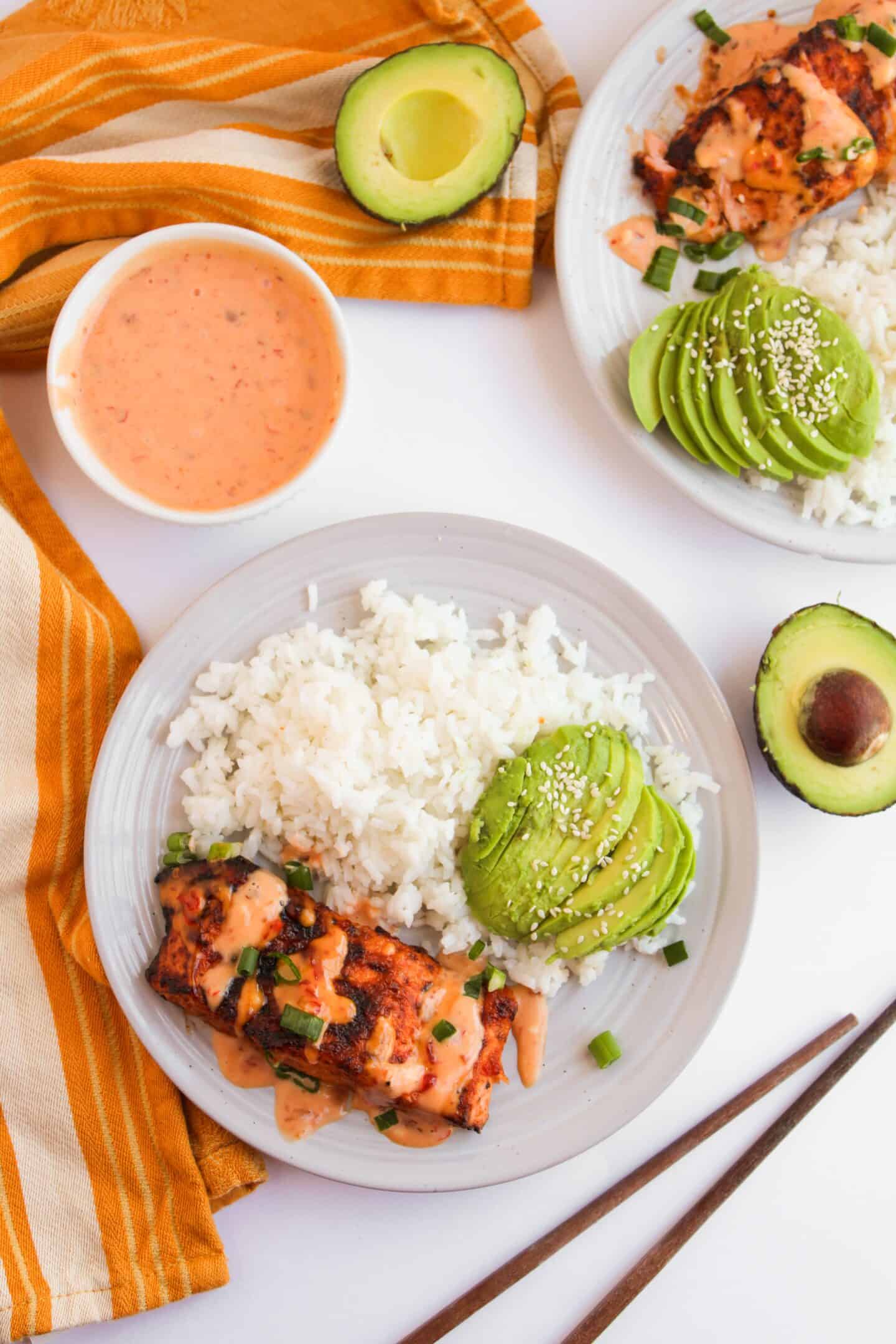 Plate with grilled chicken, white rice, and sliced avocado topped with sesame seeds. Accompanied by an avocado half, a bowl of orange sauce reminiscent of a classic seafood dish, yellow-striped cloth, and chopsticks.