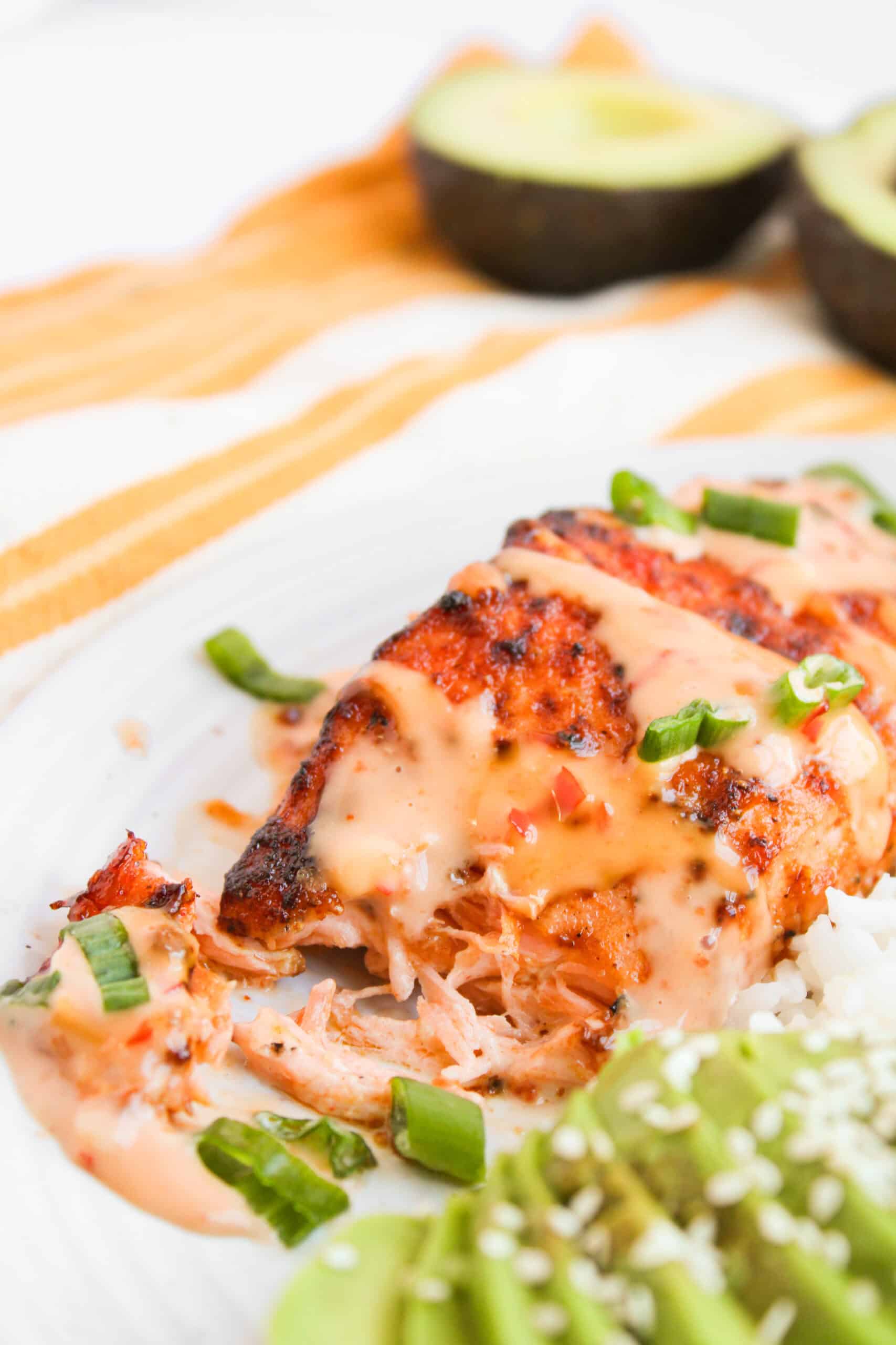 Grilled chicken with creamy sauce, sliced avocado, rice, and green onion on a white plate. In the backdrop, a halved avocado rests beside thoughts of Bang Bang Sauce dancing in your mind.