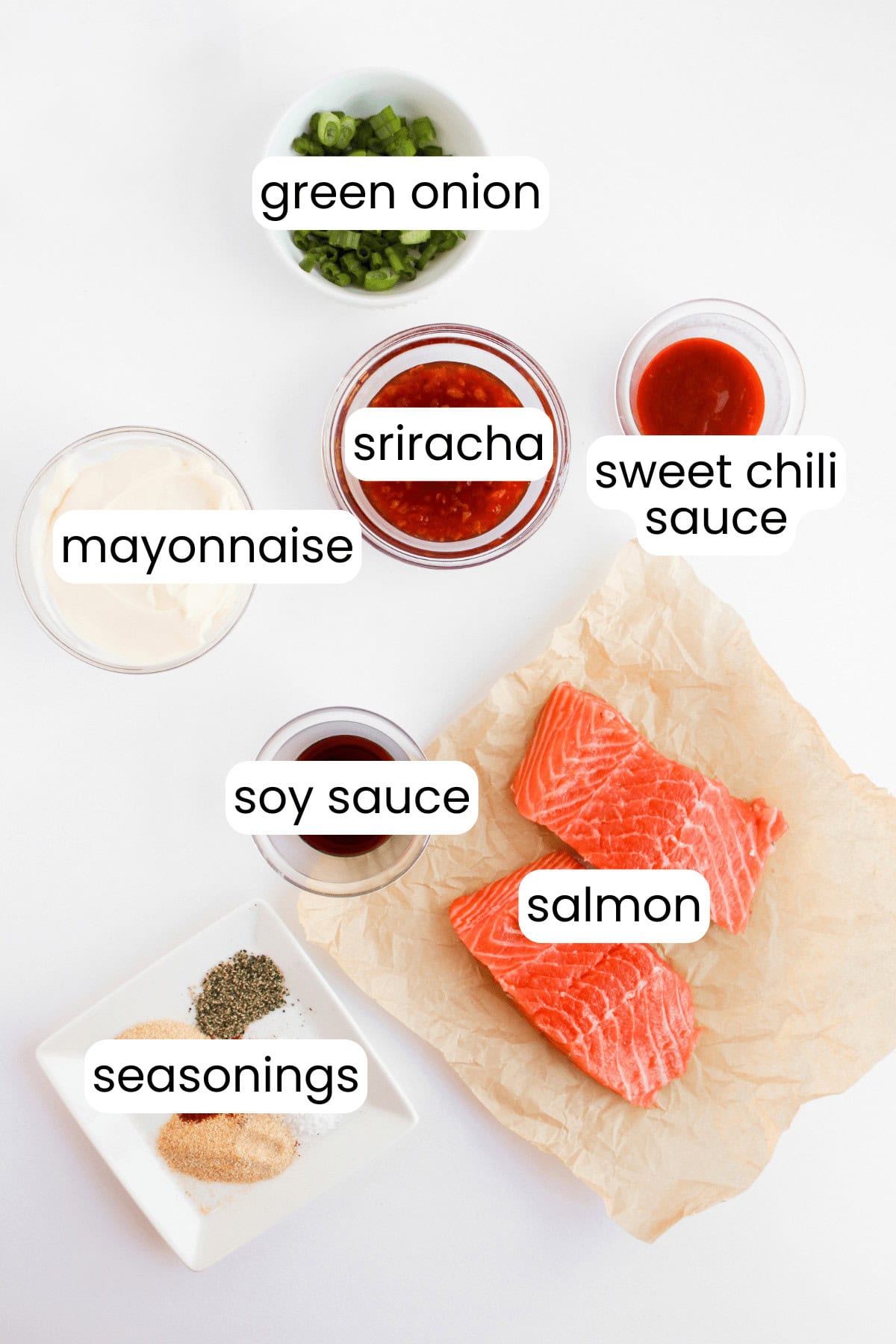 Ingredients for Bang Bang Salmon: green onion, sriracha, sweet chili sauce, mayonnaise, soy sauce, salmon, and seasonings arranged on a white surface.
