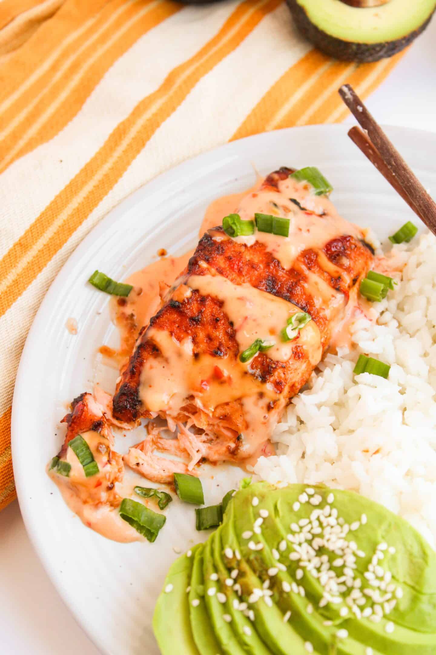 Enjoy our Bang Bang Salmon, grilled to perfection and served with white rice. Accompanied by sliced avocado topped with sesame seeds and garnished with chopped green onions, this seafood dish is beautifully presented on a white plate with chopsticks.
