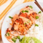 Enjoy our Bang Bang Salmon, grilled to perfection and served with white rice. Accompanied by sliced avocado topped with sesame seeds and garnished with chopped green onions, this seafood dish is beautifully presented on a white plate with chopsticks.