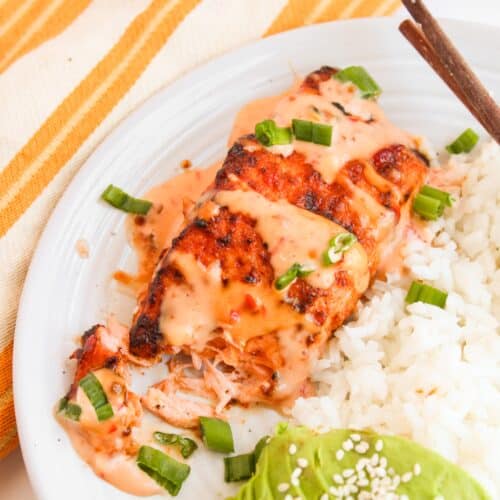 Enjoy our Bang Bang Salmon, grilled to perfection and served with white rice. Accompanied by sliced avocado topped with sesame seeds and garnished with chopped green onions, this seafood dish is beautifully presented on a white plate with chopsticks.