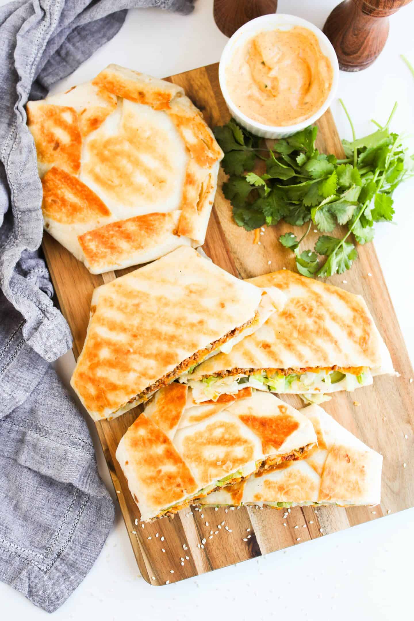 Grilled Crunchwrap halves rest on a wooden board, accompanied by a small bowl of sauce and fresh cilantro, with a gray cloth on the side.