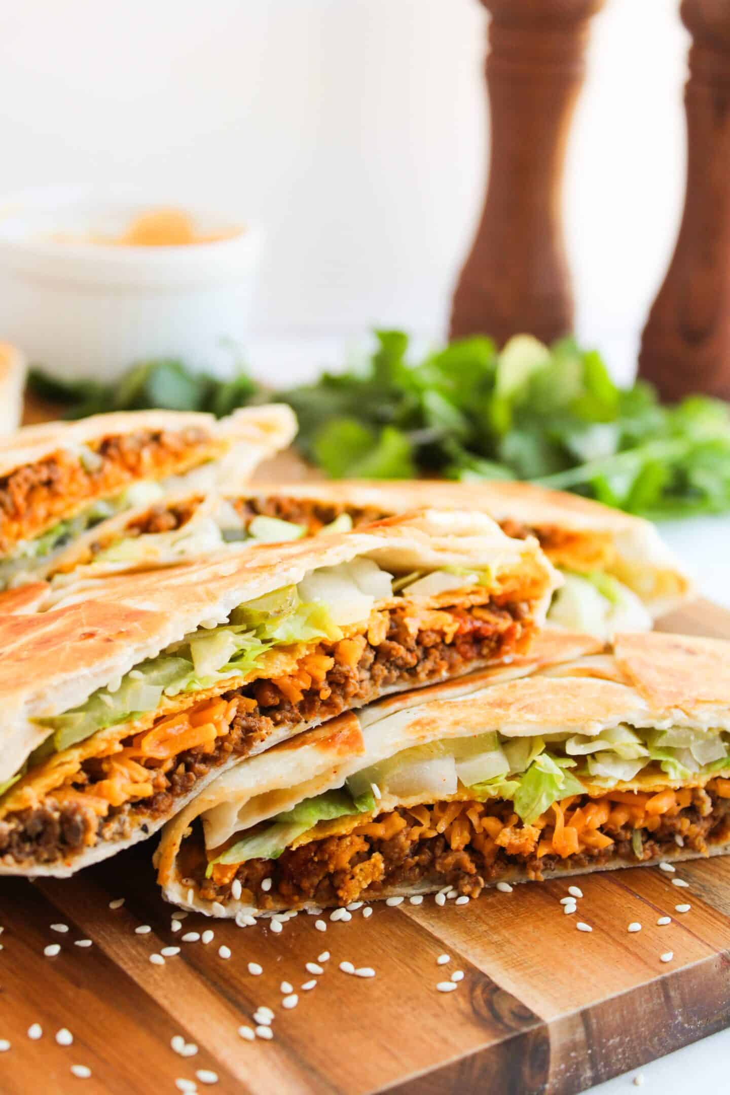 Sliced Crunchwraps filled with ground meat, lettuce, and cheese are stacked on a wooden board. Greenery and pepper grinders are in the background, creating a delightful scene reminiscent of a gourmet feast.