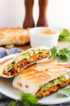 A halved, toasted Crunchwrap filled with lettuce and shredded meat rests on a white plate. A dipping sauce in a ramekin and another wrap are in the background, hinting at the zesty flavors of a Big Mac. Fresh cilantro is scattered around.