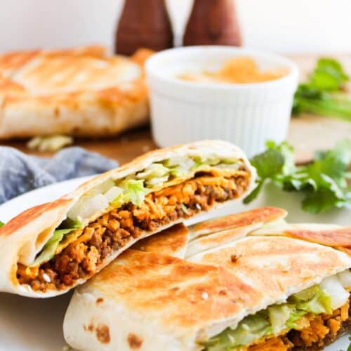 A halved, toasted Crunchwrap filled with lettuce and shredded meat rests on a white plate. A dipping sauce in a ramekin and another wrap are in the background, hinting at the zesty flavors of a Big Mac. Fresh cilantro is scattered around.