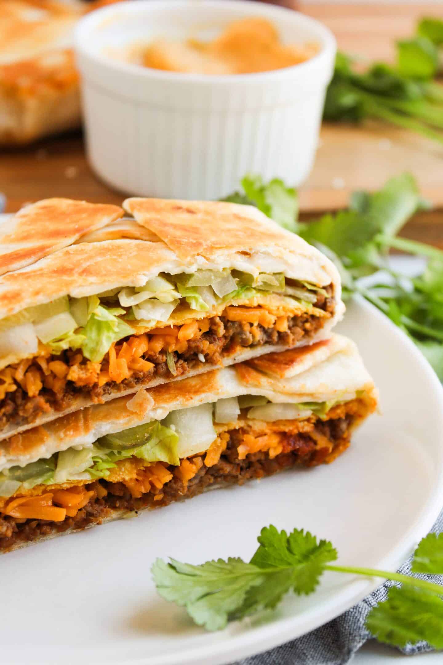 A folded, grilled Crunchwrap on a plate, filled with ground meat, lettuce, and cheese, with a side of cilantro leaves.