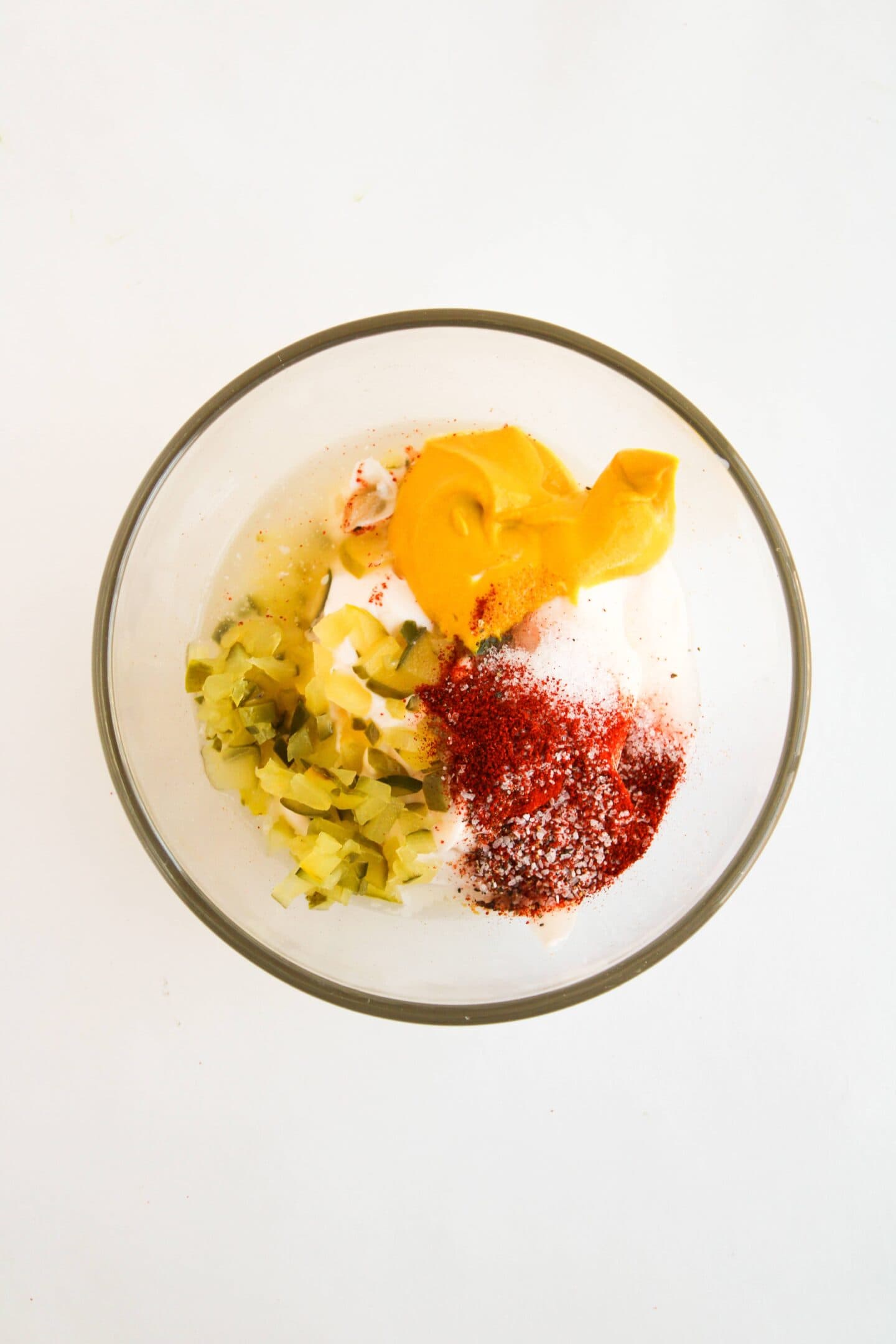 A glass bowl containing mustard, chopped pickles, mayonnaise, spices, and seasonings on a white surface evokes the classic flavors reminiscent of a Big Mac.