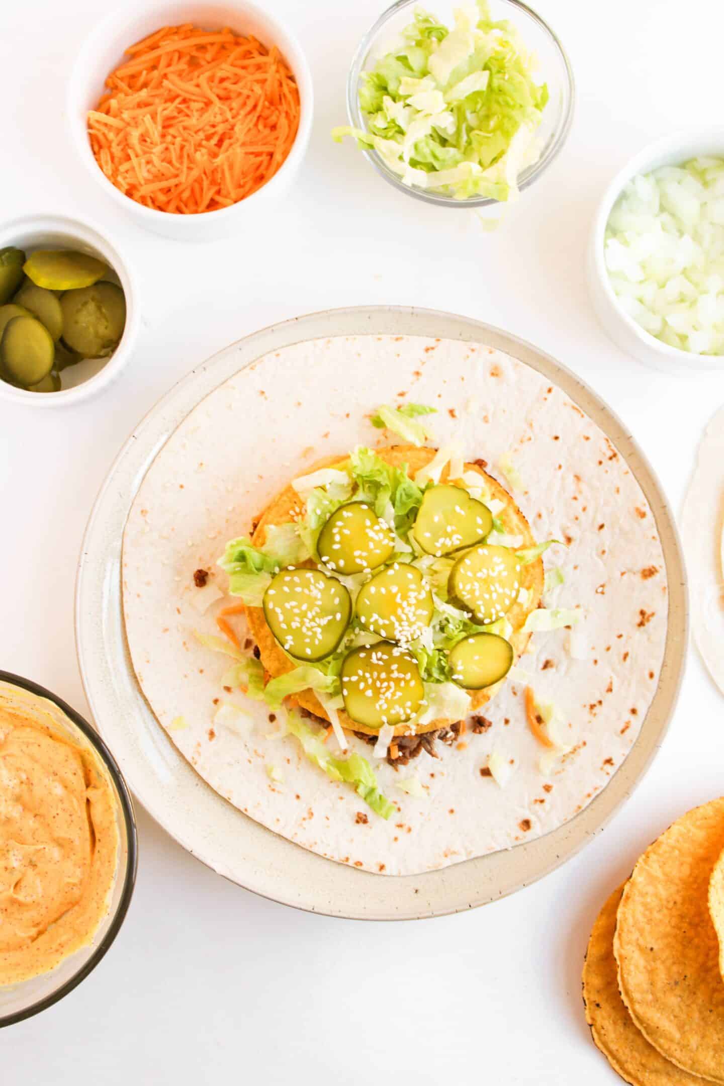 A tortilla topped with ground meat, lettuce, sliced pickles, cheese, and sesame seeds reminiscent of a Big Mac lays elegantly on a plate. Nearby are bowls of shredded carrot, extra sliced pickles, chopped onion, and a flavorful dip to complement this delightful creation.