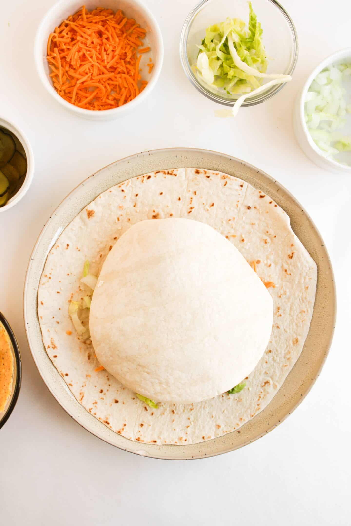 A wrap being assembled on a plate resembles a delightful Crunchwrap, with a tortilla topped with vegetables and a pita. Surrounding bowls offer grated carrots, shredded lettuce, diced onions, and sliced pickles for extra flavor.