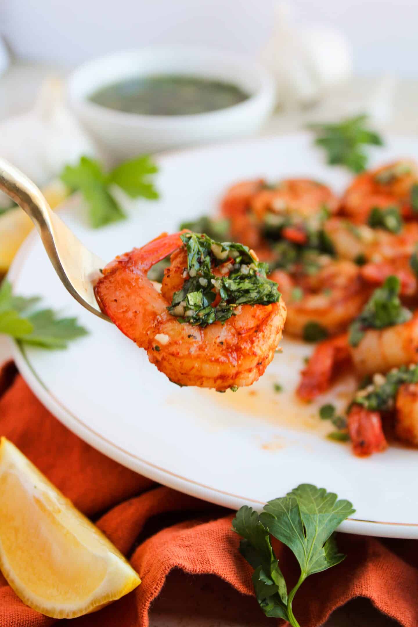 A fork holds a chimichurri-garnished shrimp, with a plate of more shrimp and parsley in the background. Lemon wedges and a sprig of parsley rest on an orange cloth, accompanied by a bowl of sauce.