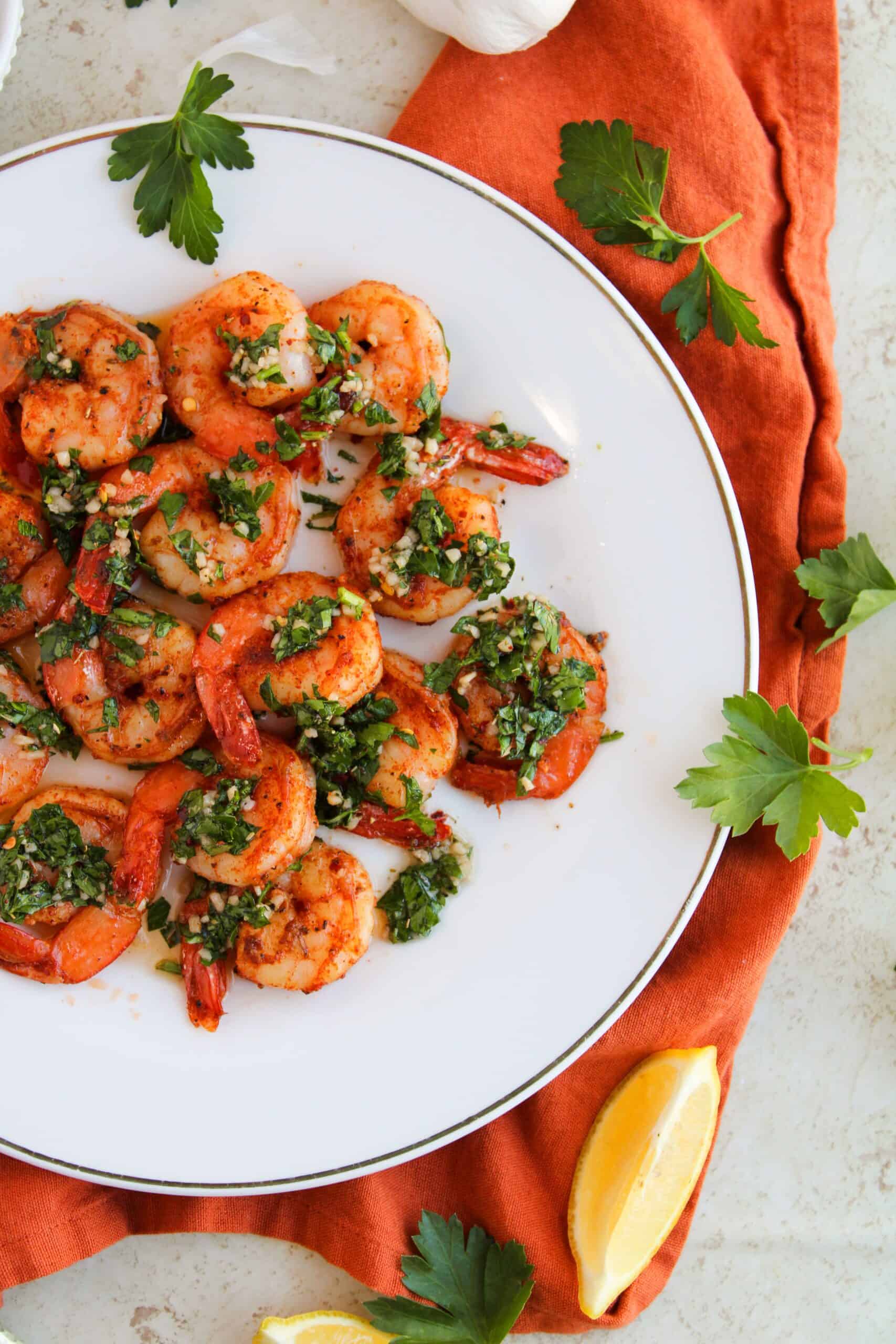 Plate of chimichurri shrimp garnished with green herbs on a white plate, set on an orange cloth with parsley and lemon wedges nearby.