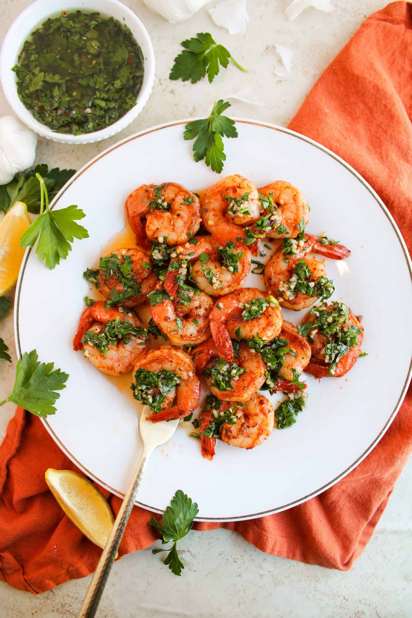 A plate of succulent shrimp topped with green herbs, served alongside a zesty chimichurri sauce and a lemon wedge, all elegantly arranged on an orange napkin.
