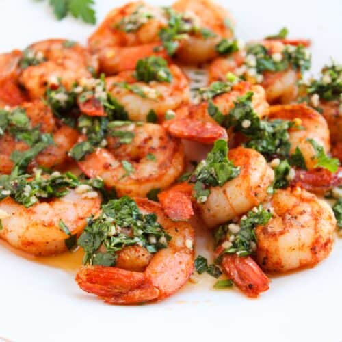 A plate of shrimp dressed in chimichurri, garnished with chopped herbs and small pieces of red pepper, sits elegantly on a white dish with an orange cloth underneath.
