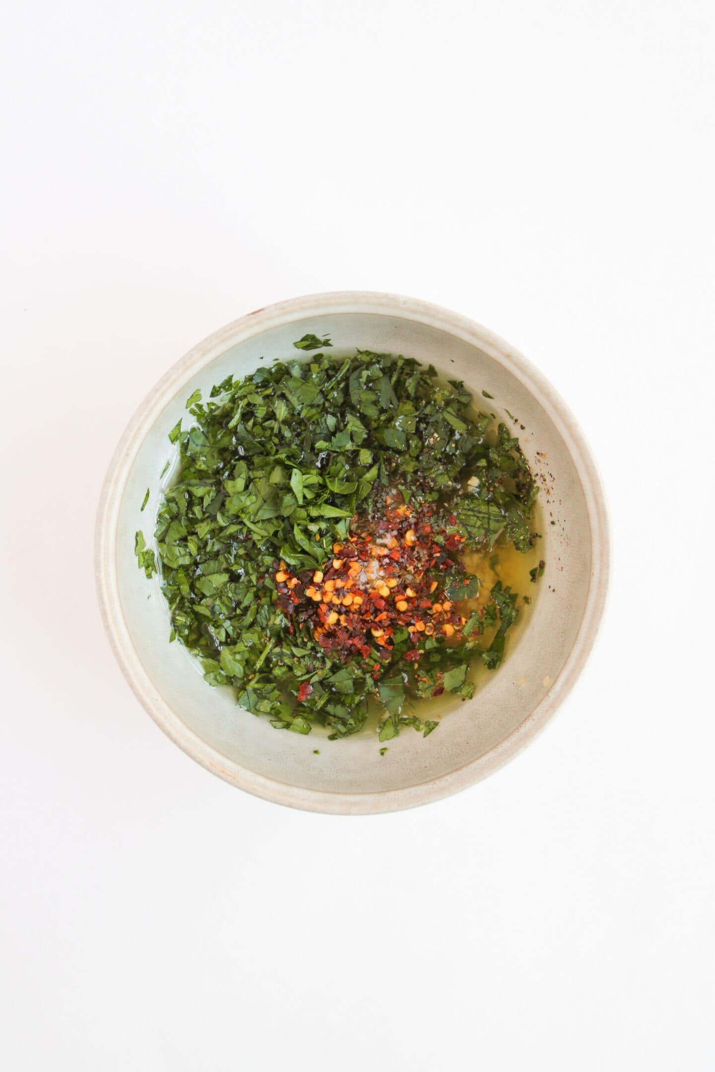 A bowl of chimichurri, a vibrant blend of chopped green herbs mixed with olive oil and red pepper flakes, rests invitingly on a white background.