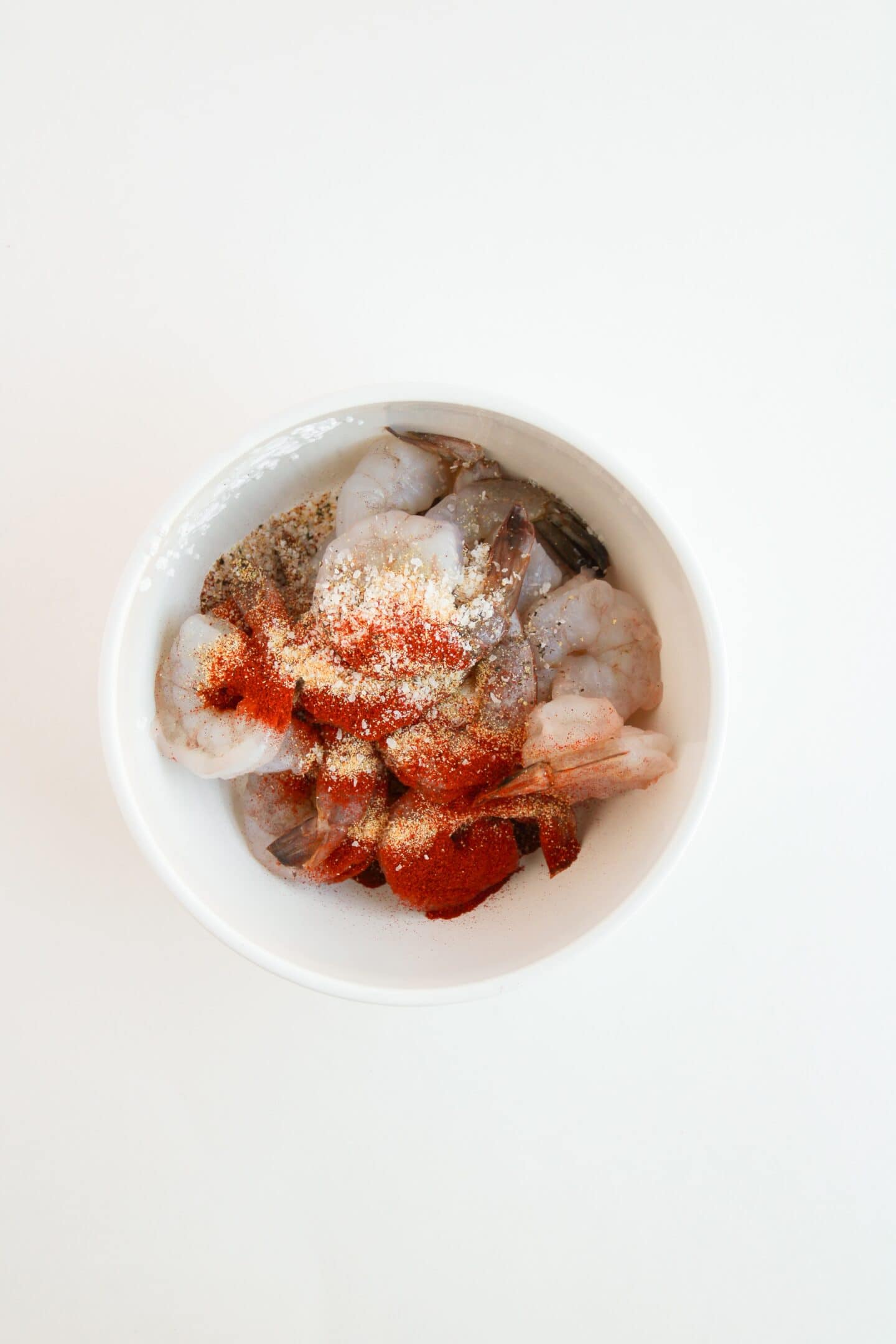 A bowl of raw shrimp is perfectly seasoned with paprika, chimichurri, and other spices on a white background.