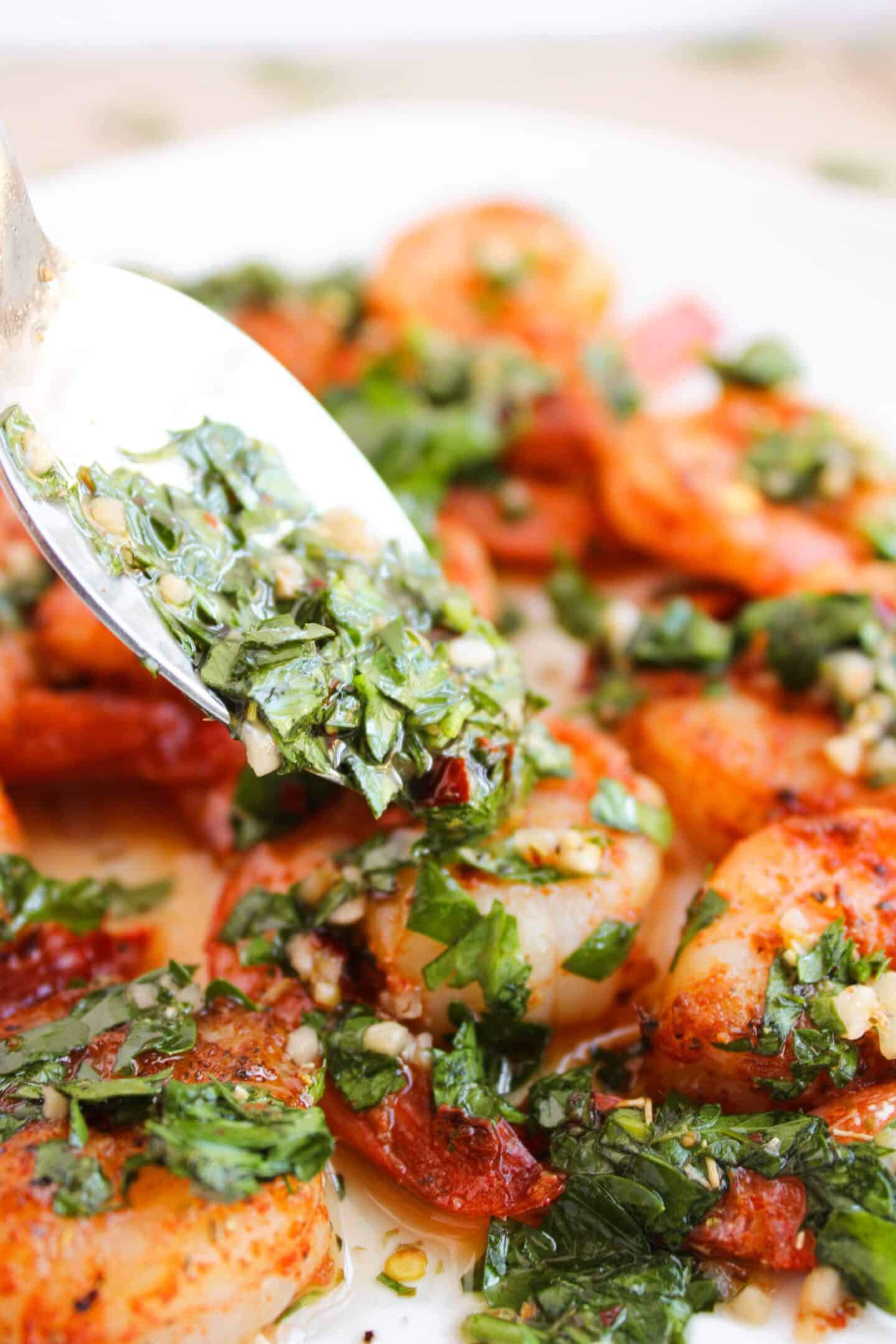 A spoon drizzles vibrant chimichurri over perfectly cooked shrimp on a plate.