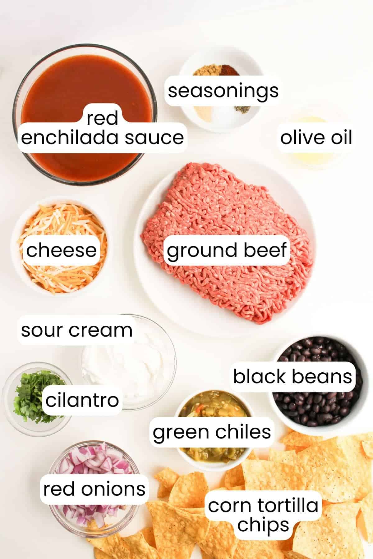 Ingredients for Beef Enchilada Nachos are artfully arranged on a sheet pan: ground beef, red enchilada sauce, shredded cheese, black beans, sour cream, green chiles, red onions, cilantro, seasonings, olive oil, and corn tortilla chips.