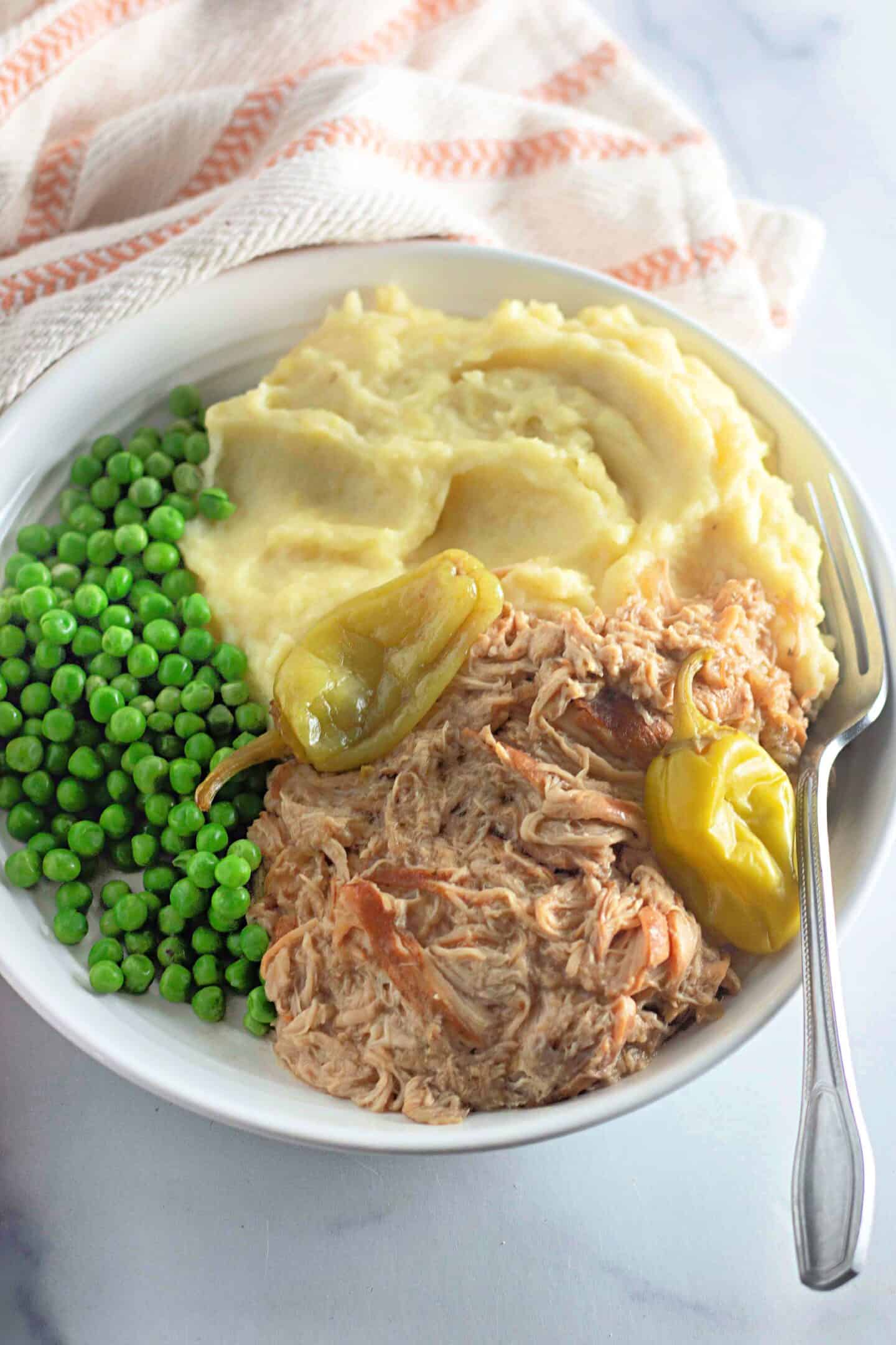 A bowl of creamy mashed potatoes, green peas, and shredded meat garnished with peppers creates a delightful Southern cuisine experience reminiscent of a Mississippi chicken recipe. A fork rests on the side, inviting you to dig in.