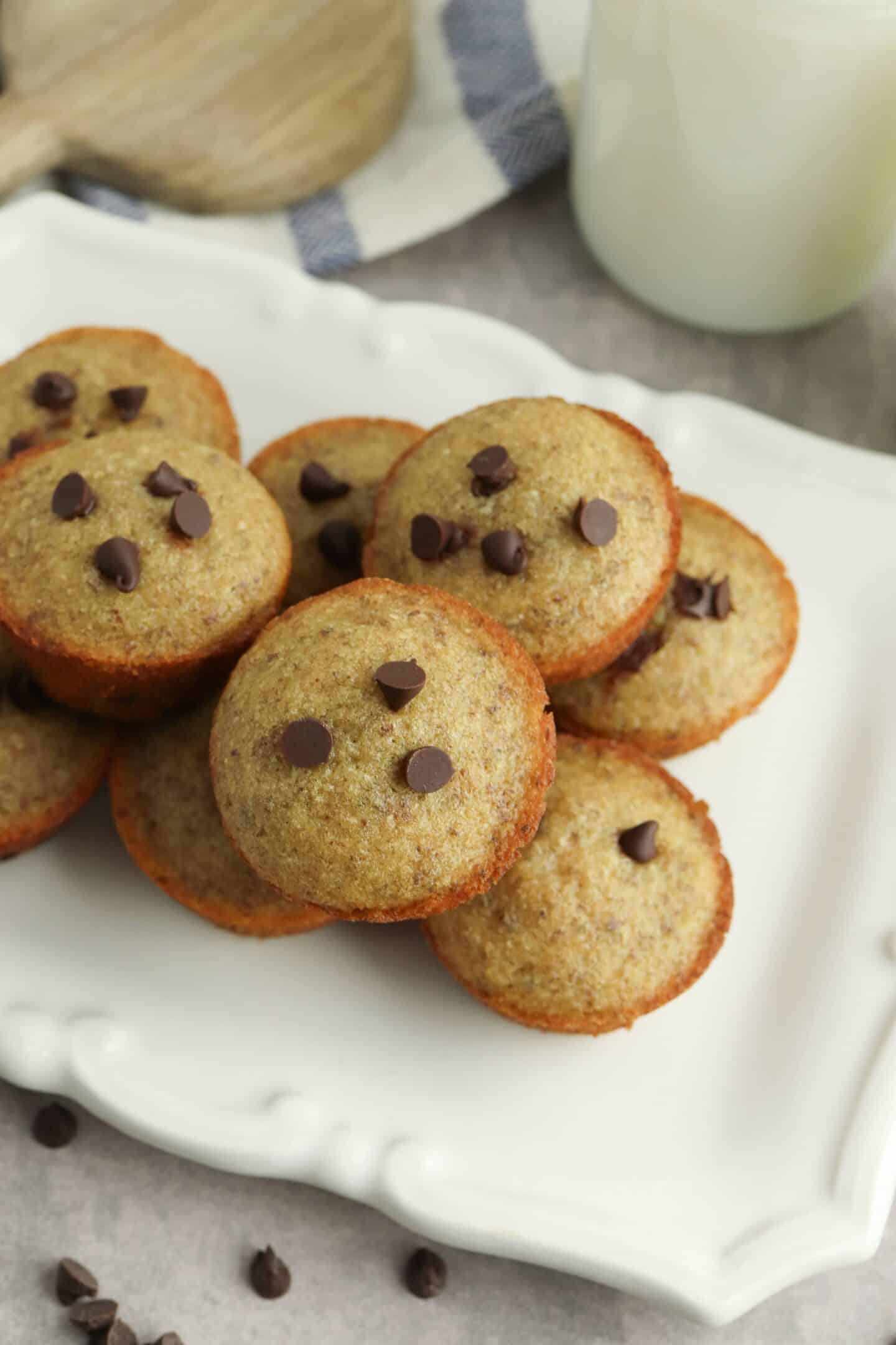 A stack of mini chocolate chip muffins is arranged on a white rectangular plate, accompanied by a glass of milk and a napkin in the background.