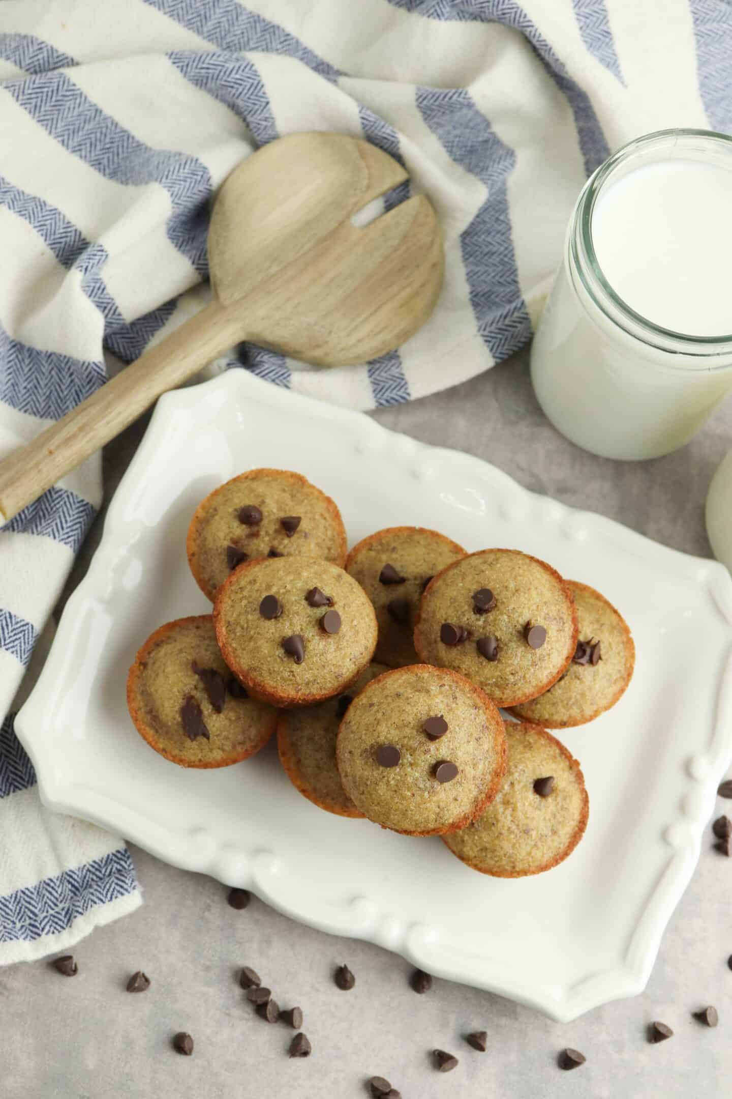 A plate of mini chocolate chip muffins is displayed, surrounded by a wooden spatula, a striped cloth, and a glass of milk.