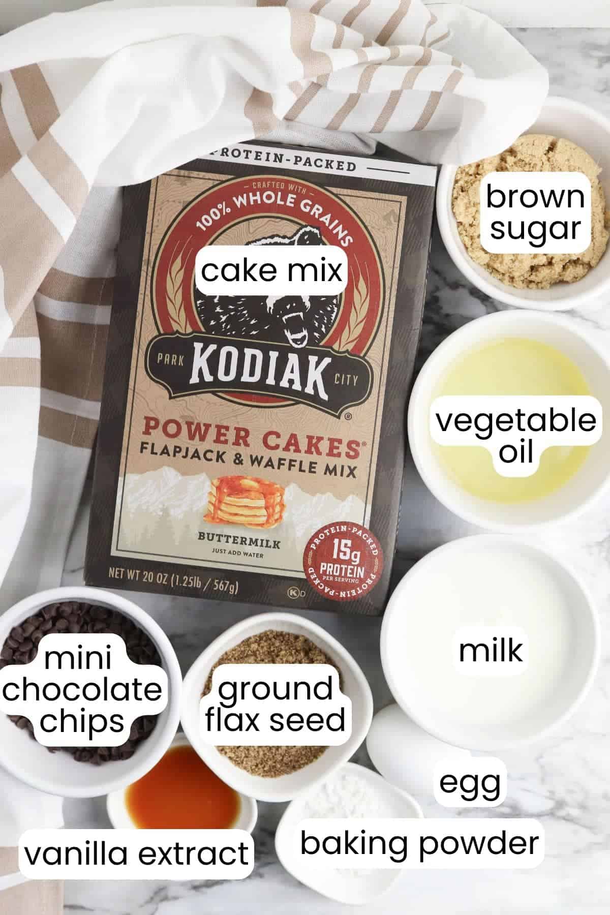 A box of Kodiak cake mix is surrounded by labeled bowls filled with brown sugar, vegetable oil, milk, egg, baking powder, vanilla extract, mini chocolate chips, and ground flax seed—perfect ingredients for protein-packed mini muffins that make deliciously healthy snacks.