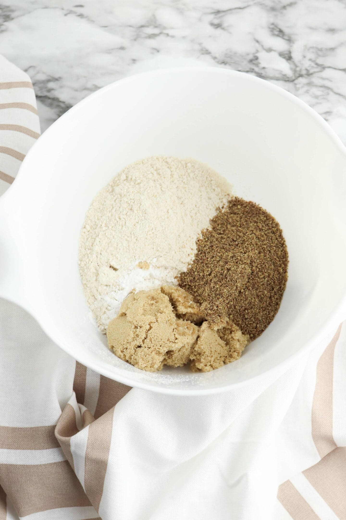 A white bowl on a striped cloth holds the perfect blend of flour, brown sugar, and ground flaxseed, ready to transform into delicious mini muffins packed with protein on a marble surface.