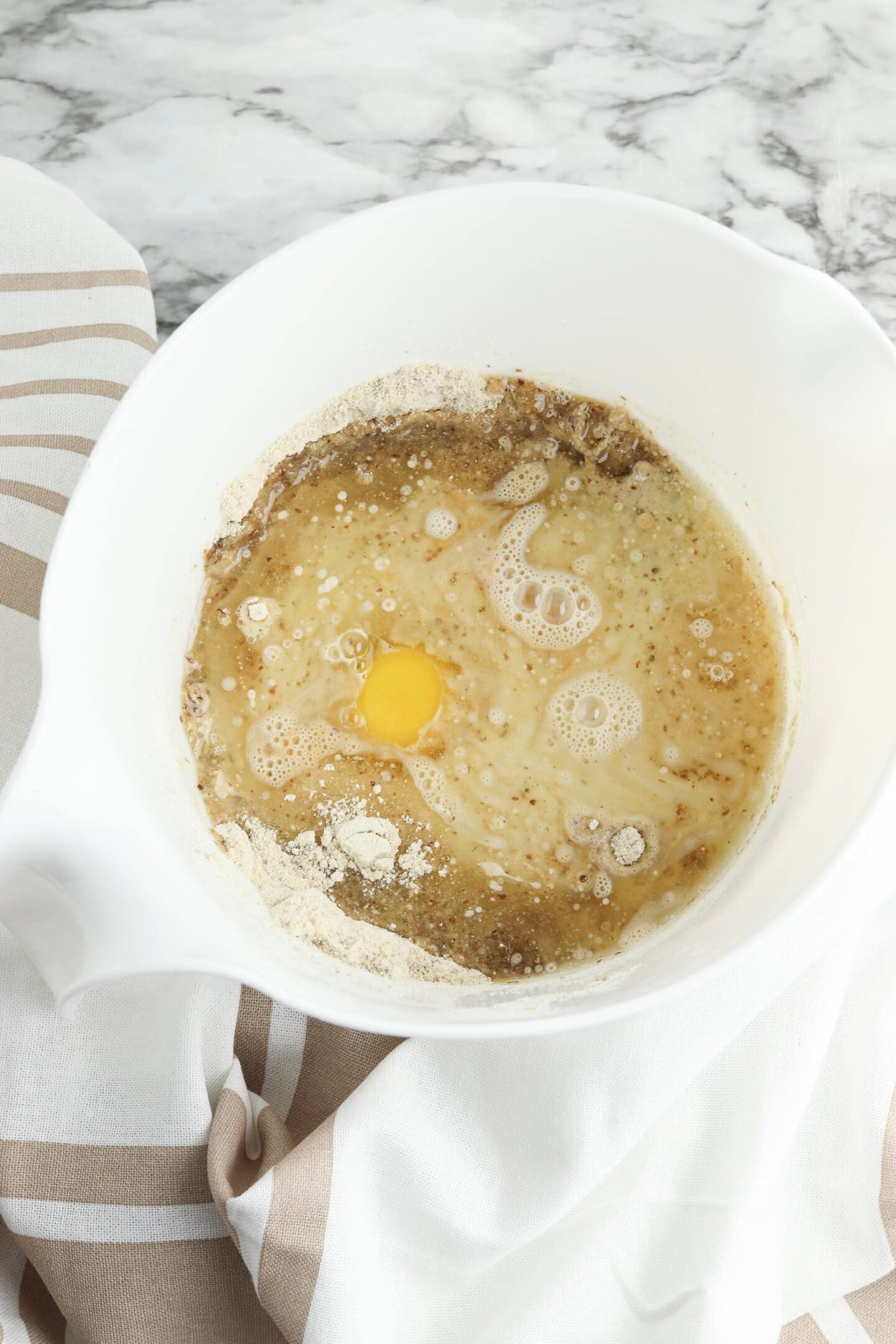 A mixing bowl with flour, a cracked egg, and a liquid rests on a marble countertop, partially covered by a striped cloth—perfect for creating protein-packed mini muffins.