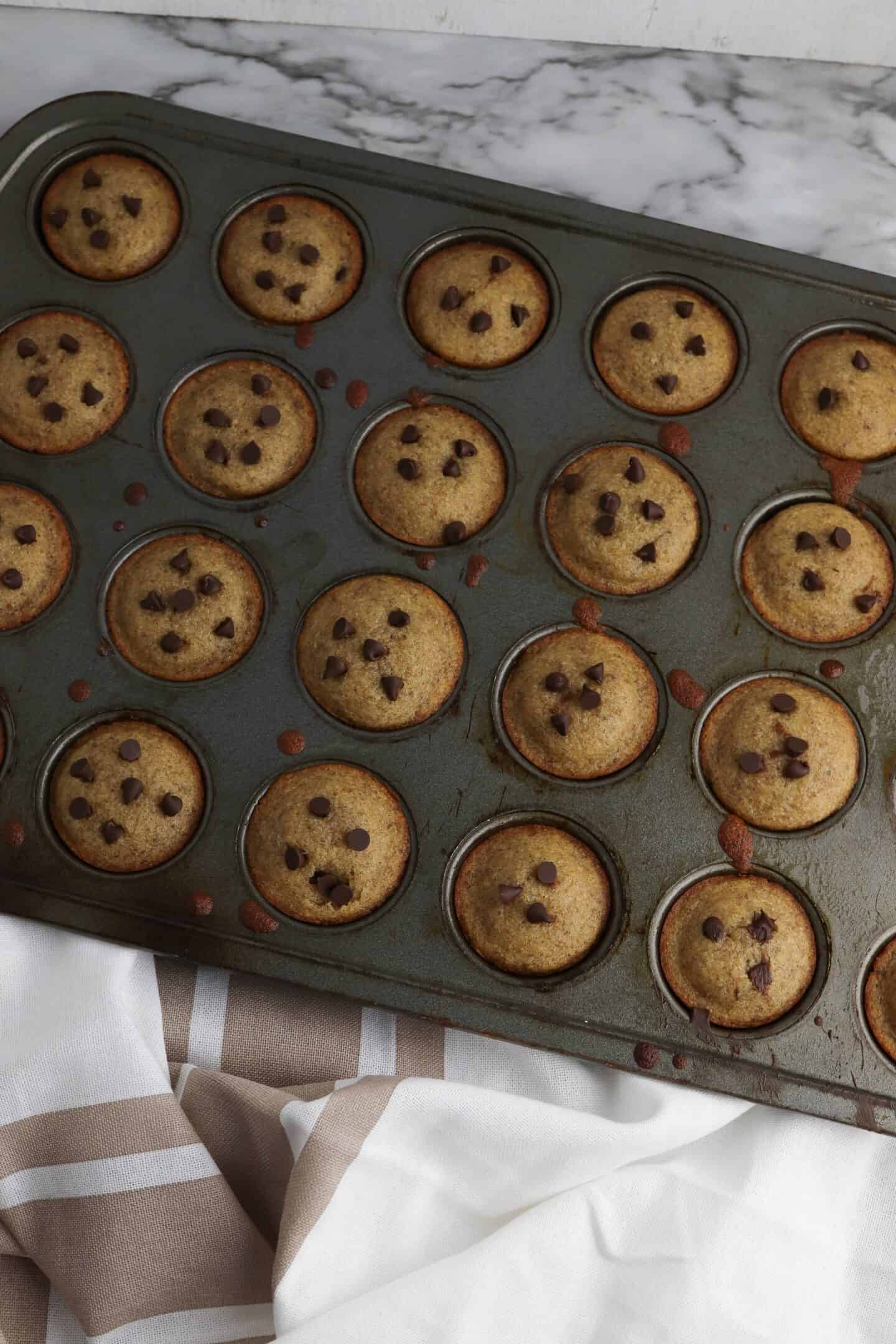 A muffin pan filled with 24 mini chocolate chip cookies rests on a marble surface, accompanied by a striped towel beneath. These delightful bites offer the perfect protein-packed treat for any occasion.