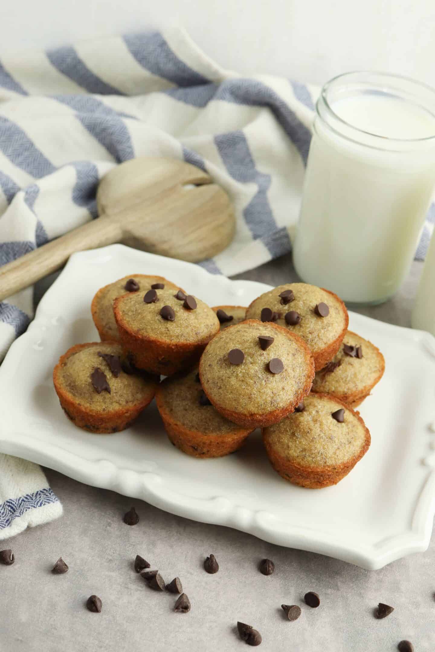 Plate of chocolate chip mini muffins on a white platter, surrounded by scattered chocolate chips. A wooden spoon, a striped cloth, and two glasses of milk rich in protein are in the background.
