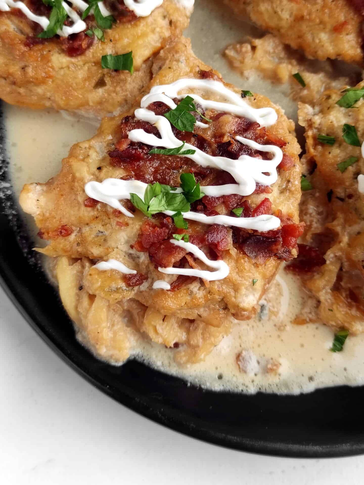 Close-up of ranch-infused chicken in creamy sauce, topped with crispy bacon bits, white sauce drizzle, and chopped parsley on a black plate.